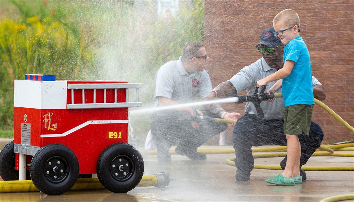 firefighter helps young boy spray fire truck target with hose