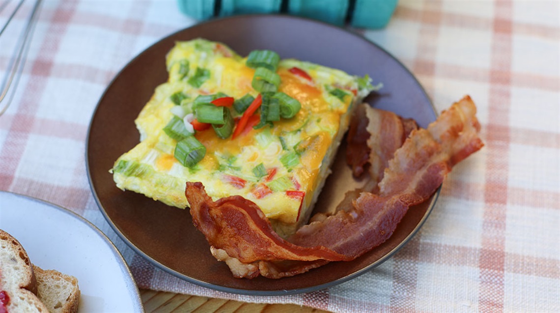 slice of breakfast casserole and bacon on plate sitting on tablecloth