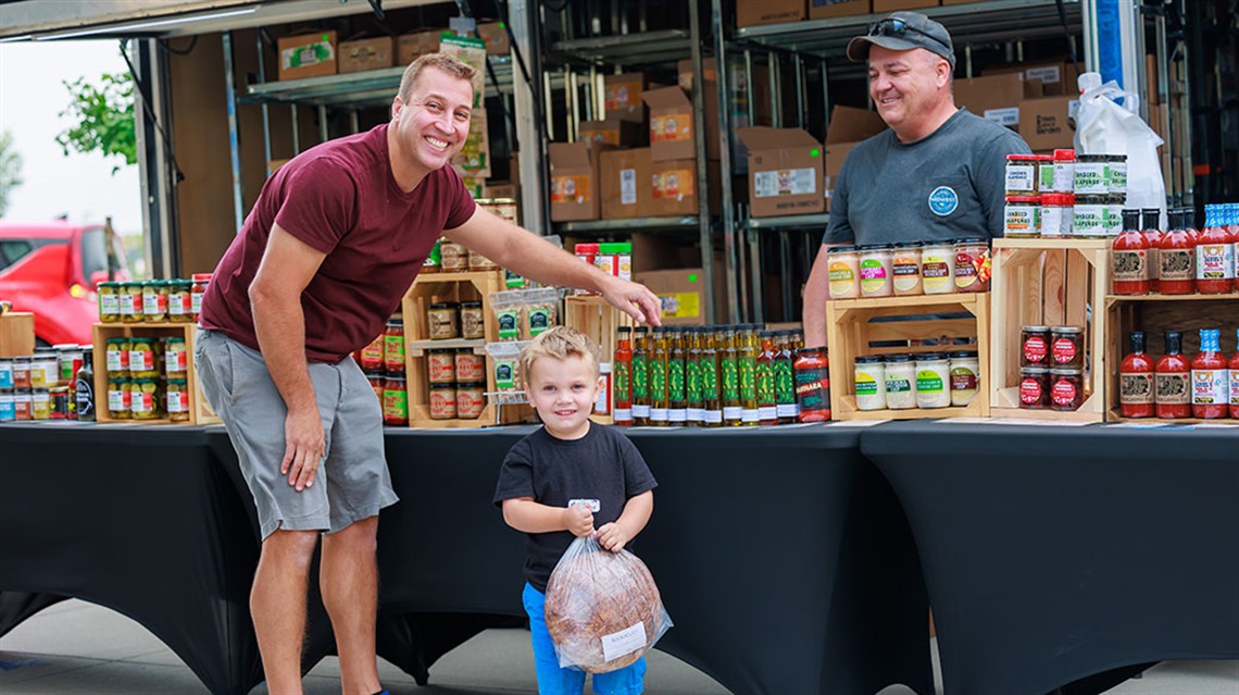 smiling dad looks at olive oils while young son holds loaf of bread at Farmers Market vendor booth