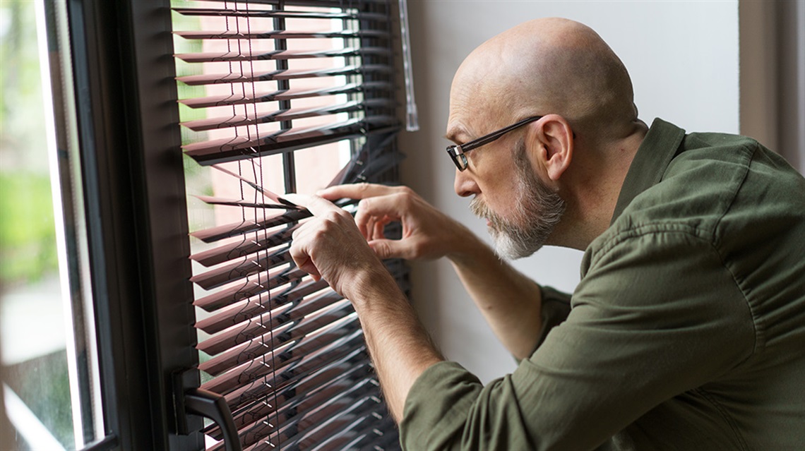 Bald man wearing glasses peers through window blinds looking outside